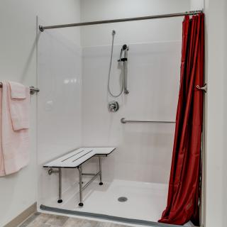 An accessible shower setup with a folding bench, handheld showerhead, grab bar, and red curtain.