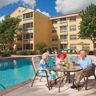 Three seniors enjoying a sunny day by the pool, sitting around a table with drinks in front of a multi-story building.