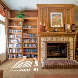 A cozy living room with a wooden bookshelf and a brick fireplace, warmly lit by sunlight streaming through the windows.