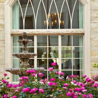 A picturesque view of a stone building's window framed by vibrant pink flowers and an ornate fountain.