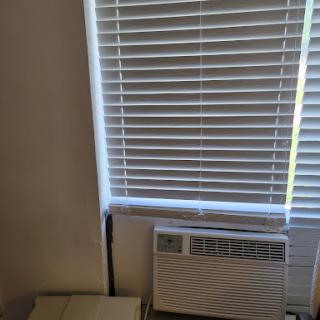 A corner of a room featuring a window with blinds and an air conditioning unit.
