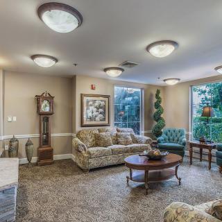 A cozy living room with traditional decor, featuring a sofa, coffee table, and a grandfather clock, enhanced by natural light from large windows.