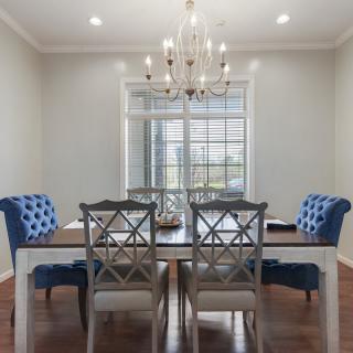 A stylish dining room features a wooden table with an elegant chandelier above, complemented by blue upholstered chairs and large windows.