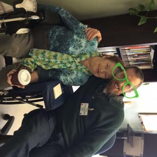 Two people sitting indoors, one wearing green novelty glasses and the other with a patterned scarf, share a fun moment.