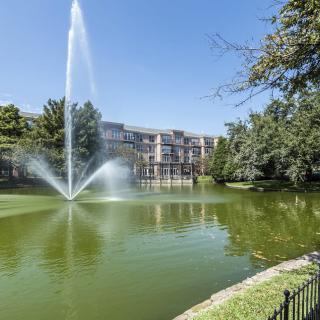 A scenic view of a fountain in a pond surrounded by trees and an apartment building.