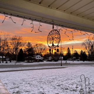 A beautiful winter sunset viewed from a snow-covered porch, with silhouettes of trees and houses in the distance.
