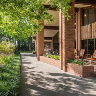 A sunny outdoor patio area with brick architecture and lush greenery along a sidewalk.