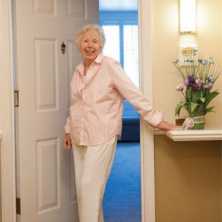 A cheerful elderly woman standing at the entrance of her home, welcoming with a smile.