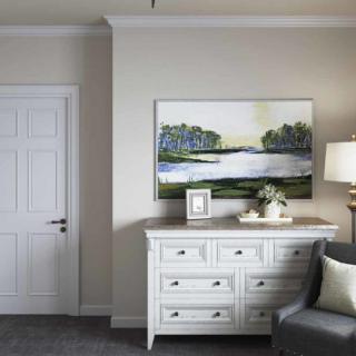 A cozy bedroom interior featuring a white dresser, artwork on the wall, and a lamp beside a comfortable chair.