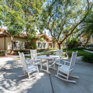 An inviting outdoor patio area with white rocking chairs and a table under shaded trees.
