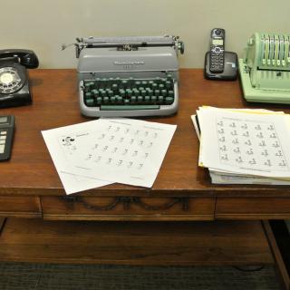 A vintage desk setup featuring a typewriter, rotary phone, calculator, and various papers.