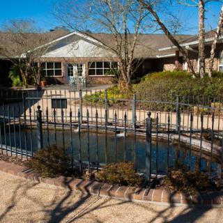 A tranquil pond surrounded by a fence in front of a building with trees under a clear blue sky.