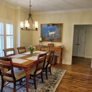A cozy dining room features a wooden table with chairs, illuminated by an elegant chandelier. A painting decorates the wall above a sideboard, adding to the warm and inviting atmosphere.