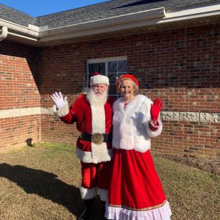 A cheerful Santa and Mrs. Claus wave outside a brick building on a sunny day.