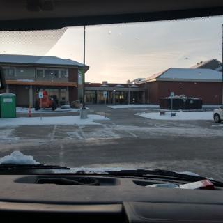 A view of a snow-covered parking area with buildings in the background.
