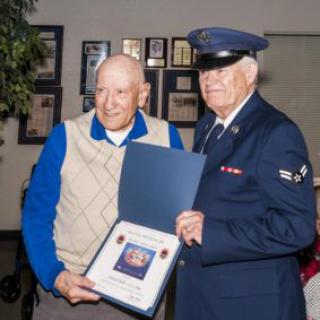 Two elderly men, one wearing a military uniform, hold a plaque during an award ceremony.