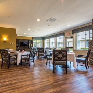 A cozy dining area with wooden floors, tables, and chairs arranged near large windows and a television.
