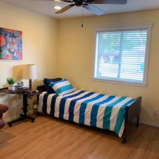 A cozy bedroom with a single bed featuring striped bedding, a desk and chair by the window, and a ceiling fan overhead.