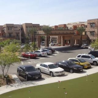 View of a parking lot in front of an apartment complex with several parked cars and surrounding greenery during daytime.