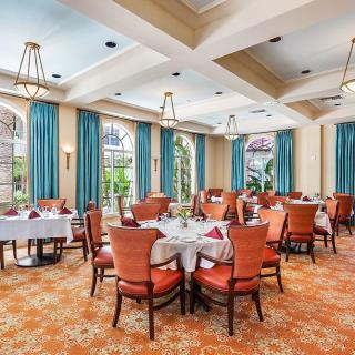 An elegant dining room with neatly arranged tables and chairs, featuring chandeliers and large windows with blue curtains.