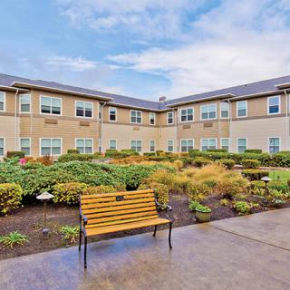 A courtyard view of a two-story building with landscaped gardens and a bench under a partly cloudy sky.