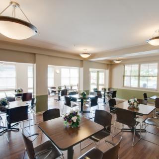 A spacious and well-lit dining area with multiple tables and chairs arranged in an organized manner, featuring large windows and overhead lighting.
