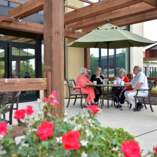 A group of people seated at a patio table under an umbrella, surrounded by flowers.