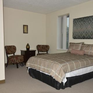 A tidy bedroom featuring a neatly made bed, two chairs with a small table in between, and minimalist decor.