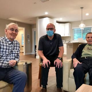 Three men sitting together in a living room setting, one wearing a mask.
