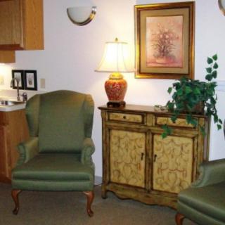 A cozy living room setup featuring green armchairs, a decorative cabinet with a lamp on top, and artwork on the wall.