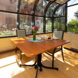A sunlit dining area features a wooden table with chairs, adorned with a vase of flowers, under a glass roof.