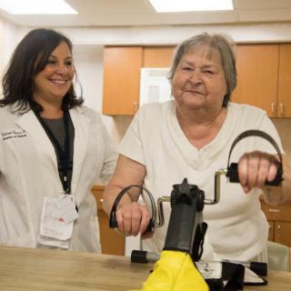 An elderly woman engages in a therapy session with a healthcare worker assisting her on exercise equipment.