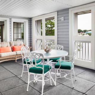 A cozy patio with a white dining set and comfortable sofa, featuring colorful cushions and ample natural light from large windows.