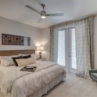 A modern bedroom with neutral colors, featuring a comfortable bed, elegant curtains, and a ceiling fan.