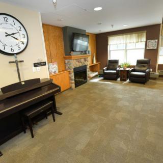 A cozy lounge area featuring a piano, large clock, fireplace, and comfortable seating with natural light streaming through the windows.