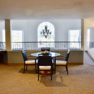 A spacious dining area with a round table and a chandelier, featuring large windows and a carpeted floor.