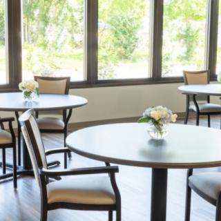 A bright dining area with round tables and chairs, featuring floral centerpieces and large windows.