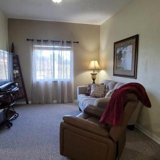 A cozy living room with a couch, recliner, and TV, warmly lit by a lamp near the window.