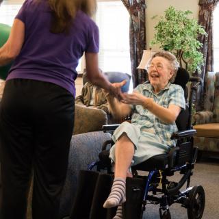 An elderly person in a wheelchair shares a joyful moment with a caregiver indoors.