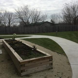 A wooden raised garden bed alongside a curved concrete pathway under an overcast sky.