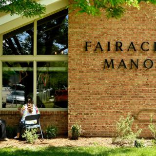 Two people sitting outside a building with a brick facade and large windows, labeled 'Fairacres Manor'.