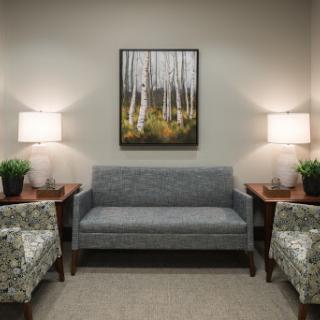 A cozy waiting area featuring a grey sofa, patterned chairs, decorative lamps, and artwork of trees.