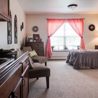 A cozy bedroom interior with a bed, dresser, and seating area, featuring natural light through bright curtains.