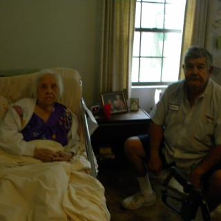 An elderly woman sitting in bed with a man seated beside her in a cozy room.