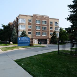A residential apartment building with a blue sign at the entrance, surrounded by trees and a driveway.