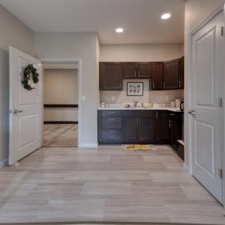 A modern apartment interior featuring a kitchen with dark cabinets, wood flooring, and an open door leading to another room.