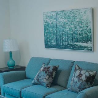 A cozy living room with a teal sofa, artistic cushions, and a matching lamp next to a forest-themed painting.