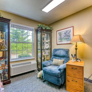 A cozy corner of a living room featuring a blue recliner chair beside a wooden side table with a lamp, surrounded by two glass cabinets displaying decorative items and an indoor plant near the window.