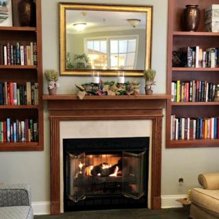 A cozy living room with a fireplace, surrounded by bookshelves and adorned with a mirror above.