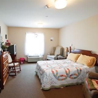 A cozy bedroom with a large bed, armchair, television, and dresser. The room is illuminated by natural light coming through the window.